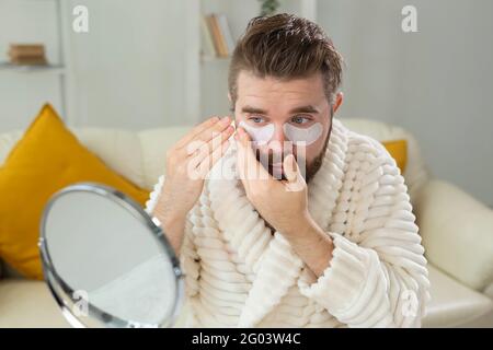 Uomo bearded che applica le macchie dell'occhio sulla sua faccia. Rughe e cura della faccia a casa per gli uomini. Foto Stock