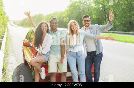 Ritratto di allegri amici multietnici che posano vicino alla loro auto a bordo strada, sorridendo a macchina fotografica, panorama Foto Stock