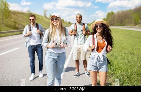 Gruppo di giovani amici diversi con zaini che camminano lungo l'autostrada, viaggiando insieme all'esterno Foto Stock