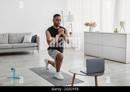Afroamericano Guy facendo Forward Lunge esercizio vicino al portatile indoor Foto Stock