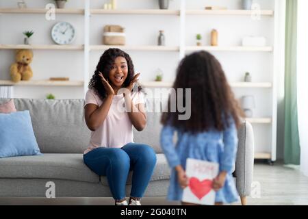 Bambina in abito nascondendo biglietto di auguri per il giorno della madre dietro la schiena, mamma eccitato felice sopra sorpresa a casa Foto Stock