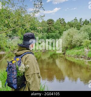 Uomo attivo in tuta protettiva con zaino in piedi vicino al fiume in una foresta. Viaggio da solo, stile di vita, concetto di turismo locale Foto Stock