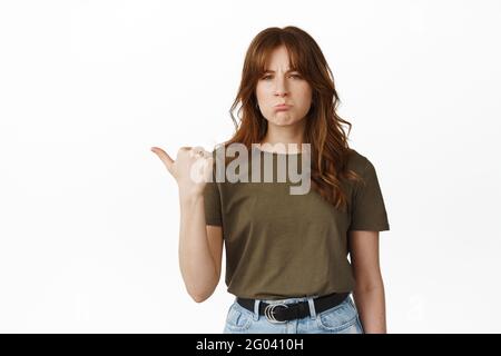 Triste e deluso studente ragazza che punta a sinistra, lamentando e sulking dispiaciuto, lamentando qualcosa, sentirsi ingiusto o rammarico, in piedi contro Foto Stock