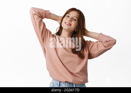 Ritratto di donna rosso spensierata che si rilassa durante il fine settimana, tenendo le mani dietro la testa e sorridendo, riposandosi e divertendosi, in piedi su sfondo bianco Foto Stock