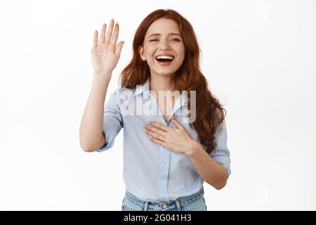 Sorridente ragazza rossa si presenta, alzando la mano e tenendo la palma sul cuore, facendo promessa, guardando felice, dica il suo nome, in piedi contro bianco Foto Stock