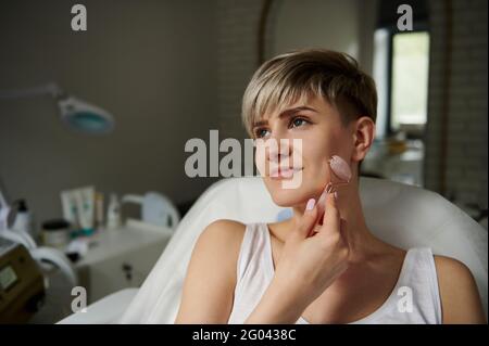 Bella e sorridente giovane donna massaggiando il viso lungo il contorno. Esecuzione di un massaggio linfodrenante con un massaggiatore a rullo con jad Foto Stock