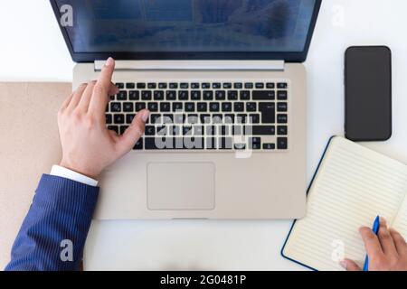 Posa piatta, mano di uomo d'affari che scrive su notebook con computer portatile e cellulare, raccoglitore e penna sulla scrivania in ufficio. Colpo di testa Foto Stock