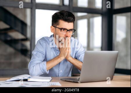 Concentrato bello giovane uomo d'affari caucasico, broker o manager indossare occhiali, seduto al tavolo in ufficio, sperando di successo nel lavoro, guardando speriamo lo schermo, le mani che si uniscono Foto Stock