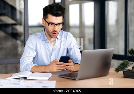 Piacevolmente scioccato uomo caucasico con, manager, broker o freelance, seduto in ufficio, utilizzando il telefono cellulare, sorpreso da notizie o messaggi, guardando lo schermo in stupore, strappando la bocca Foto Stock