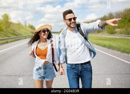 Giovane coppia allegra con zaini che camminano lungo la strada, puntando da parte, tenendo le mani, hitchhiking insieme in estate Foto Stock
