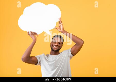 Eccitato afroamericano che tiene vuoto bolla di discorso sopra la sua testa, in posa su sfondo giallo Foto Stock