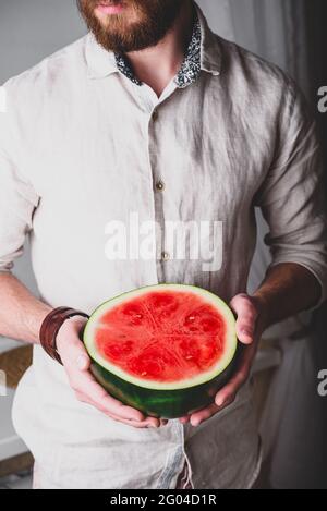 L'uomo bearded in camicia di lino tiene nelle sue mani metà di melone rosso maturo Foto Stock