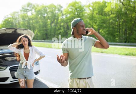 Giovane uomo che chiede aiuto con l'auto rotta, parlando al servizio di assistenza a bordo strada, la sua ragazza sconvolgente in piedi nelle vicinanze Foto Stock