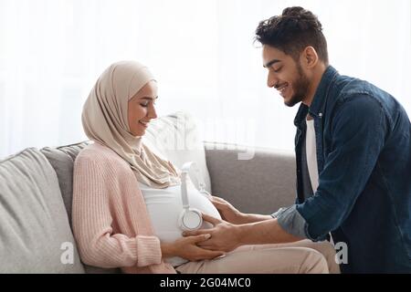 Adorabile marito arabo che mette le cuffie wireless sul venticellano della moglie incinta Foto Stock