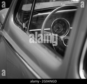 Immagine in scala di grigi del volante e del tachimetro di un'auto d'epoca visto dal finestrino laterale della portiera anteriore Foto Stock