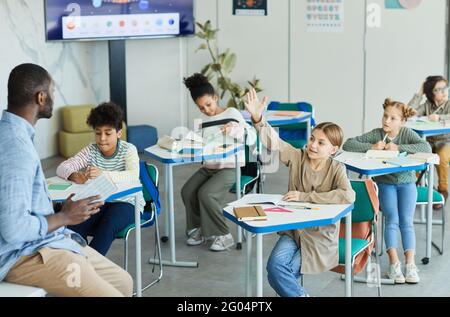 Gruppo vario di bambini in classe scolastica con ragazza sorridente che alza la mano, spazio di copia Foto Stock