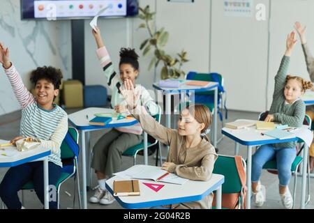 Gruppo vario di bambini felici che alzano le mani in classe scolastica, spazio di copia Foto Stock