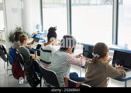 Vista posteriore a diversi gruppi di bambini seduti in fila in classe scolastica e utilizzando computer, spazio di copia Foto Stock