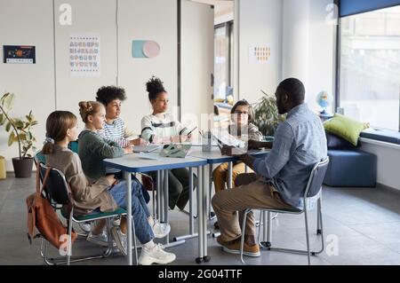 Foto a lunghezza intera di diversi gruppi di bambini seduti a. tavolo con insegnante di sesso maschile in classe moderna Foto Stock