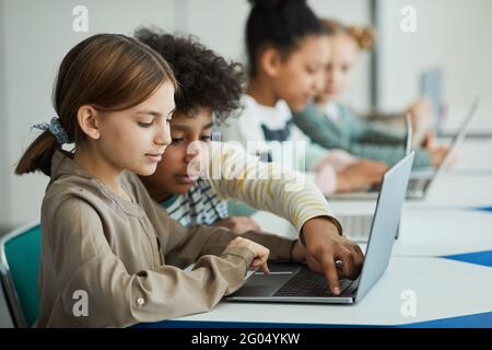 Vista laterale su diversi gruppi di bambini seduti di fila in classe scolastica e con l'uso di computer portatili Foto Stock