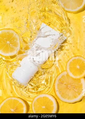 Tubo bianco vuoto cosmetico in acqua trasparente pulita con fette di limone su sfondo giallo. Spruzzi d'acqua sulla superficie, luce solare. Appiattita verticale, spazio di copia. Mockup di bellezza per crema o idratante Foto Stock