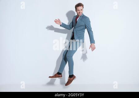 immagine completa del corpo di un uomo d'affari felice bearded in un vestito elegante sorridente e saltando nell'aria mentre si diverte e si posa su sfondo grigio in s. Foto Stock