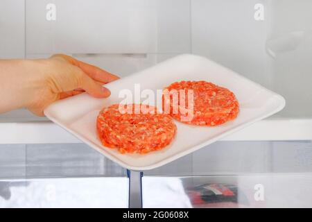 Il Burger di pollo crudo in contenitore è su ripiano in frigorifero aperto. La mano femminile tira fuori dal frigorifero un ingrediente per cucinare piatti di carne. Foto Stock