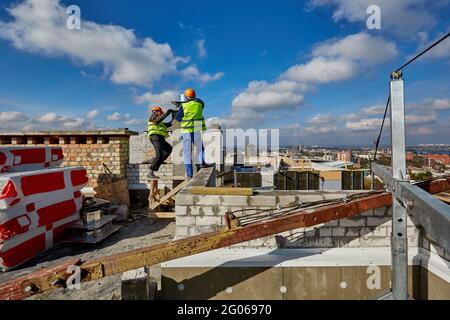 Due lavoratori in abbigliamento protettivo e caschi di sicurezza lavorano con un sistema di ventilazione sul tetto dell'edificio in costruzione Foto Stock