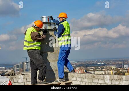 Due lavoratori in abbigliamento protettivo e caschi di sicurezza stanno installando un tubo per il sistema di ventilazione sul tetto di un edificio moderno in costruzione Foto Stock