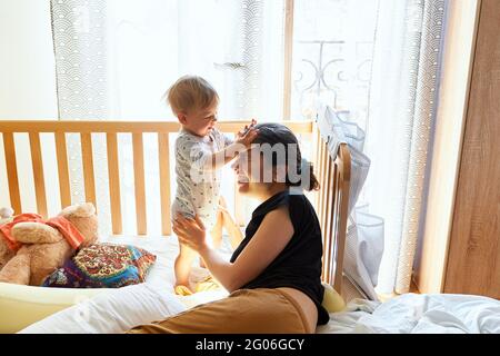 Il ragazzino gli aggrappa le mani sulla fronte della madre sorridente che giace sul letto Foto Stock