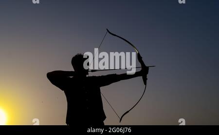 un uomo dalla silhouette che tiene un arco e una freccia davanti al tramonto. Foto Stock