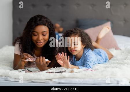 Felice madre nera e la sua bella figlia facendo manicure, limando e dipingendo le loro unghie, sdraiati sul letto a casa Foto Stock