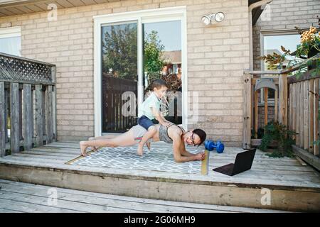 Attività sportive per la famiglia. Madre caucasica di mezza età con il figlio del toddler che fa l'allenamento in linea remoto lontano di idoneità sul cortile della casa con il laptop. Genitore Foto Stock