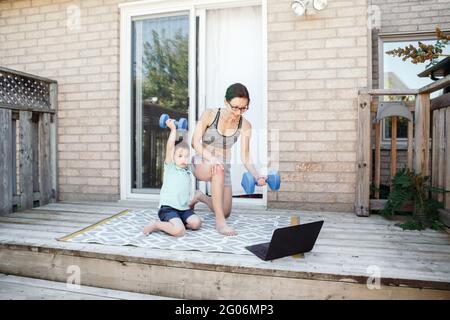 Attività sportive per la famiglia. Madre caucasica di mezza età con il figlio del toddler che fa l'allenamento remoto in linea di idoneità lontano con le campane-dumb sul cortile con il lapto Foto Stock
