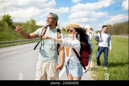 Viaggiatori millenari con zaini a piedi lungo la strada, escursioni in campagna. Gruppo di persone che hanno un viaggio autoststop Foto Stock