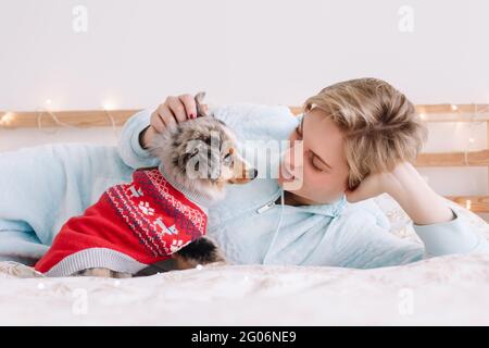 Giovane donna caucasica con capelli corti sdraiati a letto con simpatico cane cucciolo a casa. Il proprietario del PET accarezza il piccolo amico di furry. Rapporto di domest Foto Stock