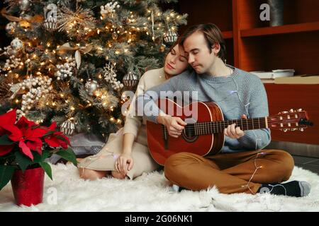 Uomo e donna festeggiano insieme Natale o Capodanno. Uomo che suona la chitarra e canta per donna. Giovane coppia che festeggia le vacanze invernali t Foto Stock