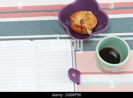 Per fare l'elenco in blocco note con caffè e dolci sul tavolo Foto Stock