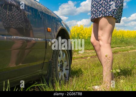 Gambe slanciate di una giovane donna in una gonna stand accanto a una macchina sporca bloccato su una strada sterrata nel mezzo di un campo di colza Foto Stock