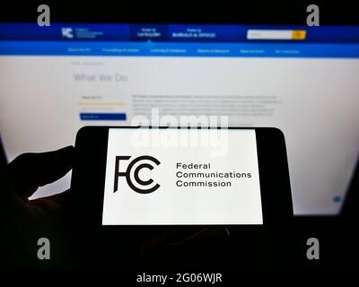 Persona che detiene il telefono cellulare con il sigillo della Federal Communications Commission (FCC) dell'agenzia statunitense sullo schermo davanti alla pagina web. Mettere a fuoco il display del telefono. Foto Stock