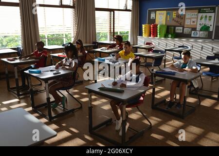 Gruppo diversificato di studenti che studiano mentre si siedono sulla loro scrivania in classe alla scuola elementare Foto Stock