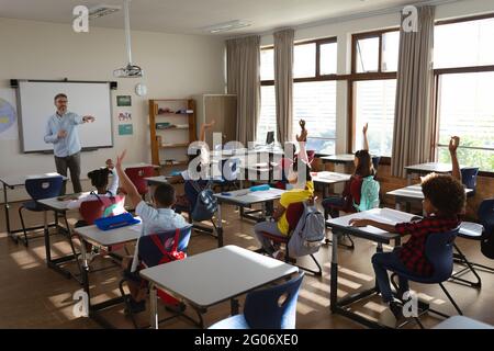 Gruppo di studenti diversi che alzano le mani nella classe delle scuole elementari Foto Stock