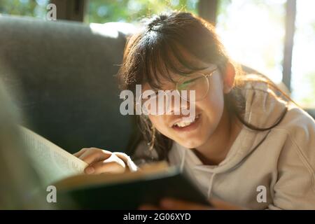 Sorridente ragazza asiatica in occhiali leggere un libro e sdraiato sul divano Foto Stock