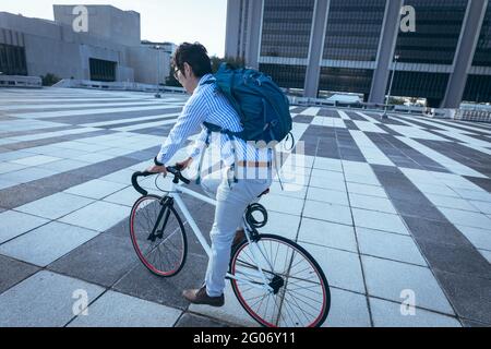 Uomo d'affari asiatico in bicicletta in strada con edifici moderni sullo sfondo Foto Stock