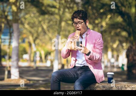 Uomo d'affari asiatico che utilizza smartphone bere caffè da asporto attraversando la strada della città Foto Stock