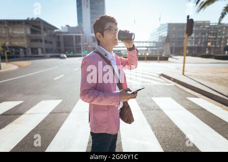 Uomo d'affari asiatico che utilizza smartphone bere caffè da asporto attraversando la strada della città Foto Stock