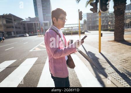 Uomo d'affari asiatico che utilizza smartphone bere caffè da asporto attraversando la strada della città Foto Stock