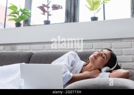 giovane donna adulta tranquilla con gli occhi chiusi in cuffie poggiate sul divano vicino al computer portatile a casa Foto Stock