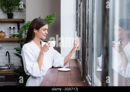 bella giovane donna adulta in accappatoio prendendo selfie sul cellulare vicino alla finestra in loft moderno Foto Stock