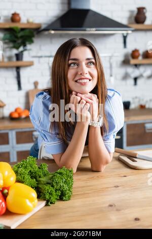 giovane donna adulta in piedi vicino al tavolo con verdure e tenendo le mani vicino al viso in cucina Foto Stock